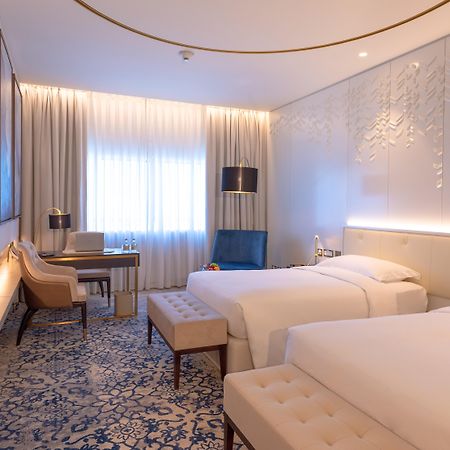 Steigenberger Hotel Doha Exterior photo The photo shows a modern hotel room with two single beds placed beside each other. The room features a contemporary design with light-colored walls and stylish decor. There is a large window dressed with sheer curtains, allowing natural light to fill