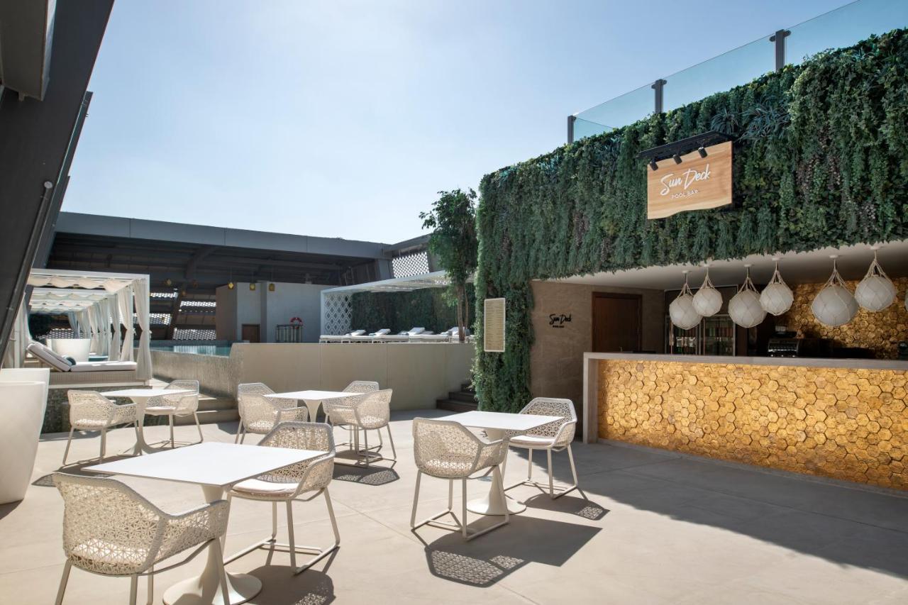Steigenberger Hotel Doha Exterior photo The photo shows a stylish outdoor seating area of a modern venue, likely a restaurant or bar. The space features several white tables and chairs arranged neatly on a light-colored floor. In the background, there's a bar area with a warm-toned wood fa