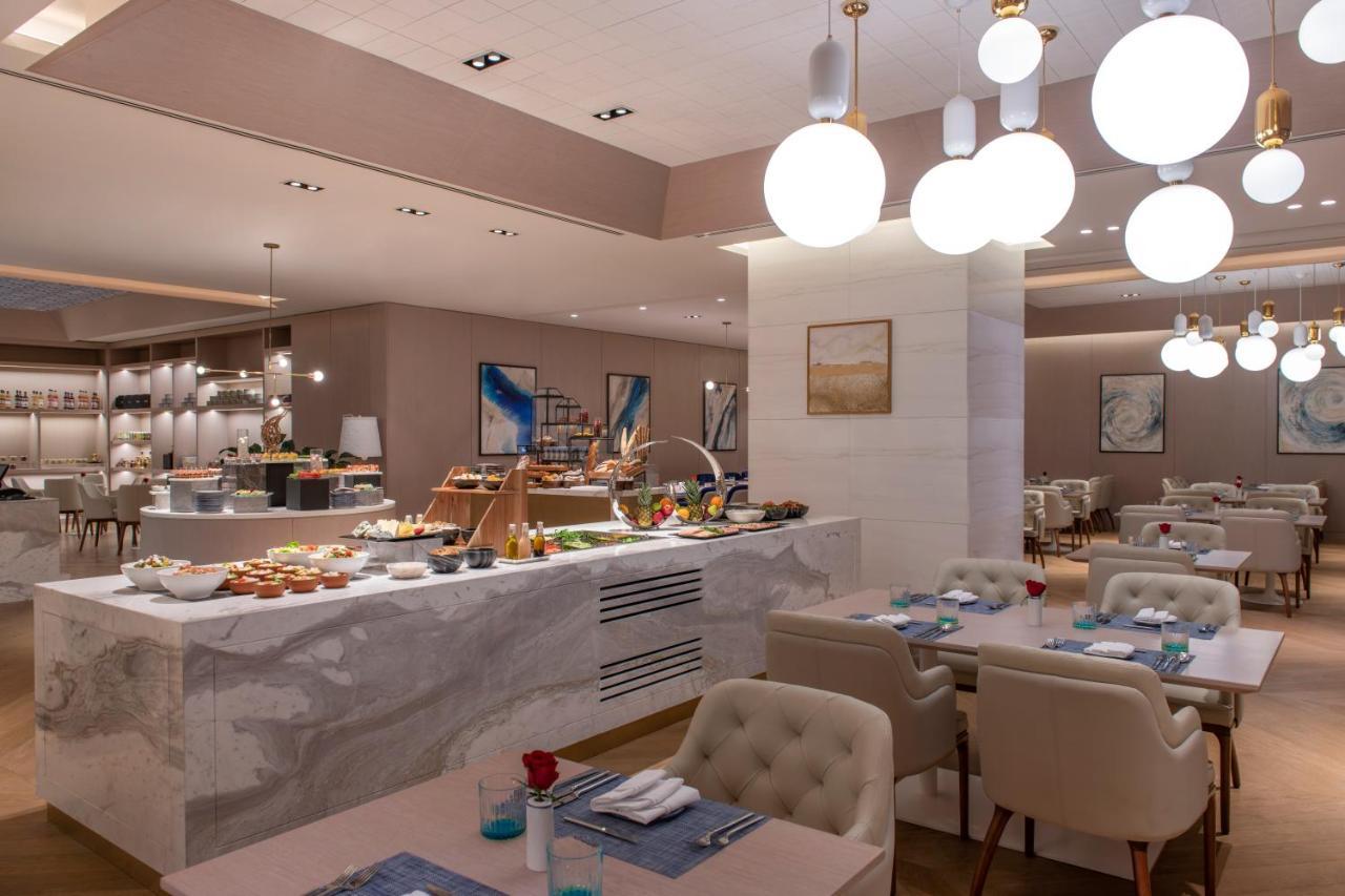 Steigenberger Hotel Doha Exterior photo The photo shows a modern restaurant interior that features a stylish and elegant design. In the foreground, there are tables with light-colored upholstered chairs, set with blue napkins and small flower arrangements. 

In the background, there is a b