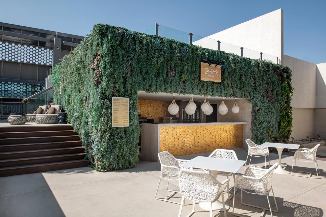 Steigenberger Hotel Doha Exterior photo The photo shows an outdoor bar area surrounded by lush greenery. The bar has a modern design, featuring a wooden counter with decorative pendant lights hanging above. There are several white outdoor tables and chairs arranged in front of the bar, whi
