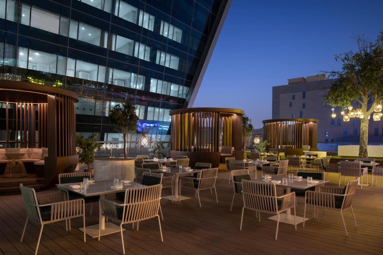 Steigenberger Hotel Doha Exterior photo The photo shows an outdoor dining area that features several tables and chairs arranged neatly on a wooden deck. The seating includes modern white chairs and a mix of seating styles, including some areas with privacy screens or structures that provid