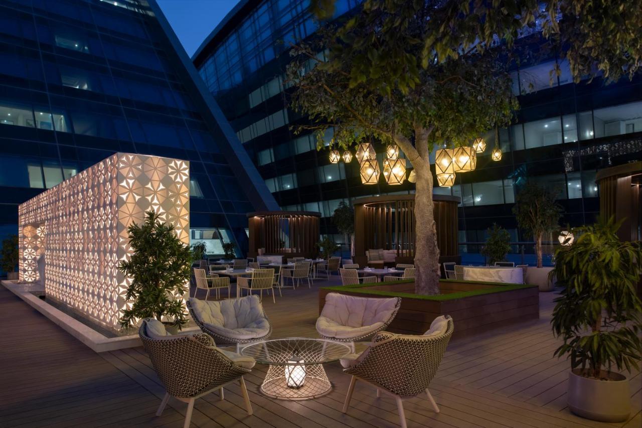 Steigenberger Hotel Doha Exterior photo The photo depicts a stylish outdoor terrace area at night. There are modern seating arrangements, featuring woven chairs and a decorative table, surrounded by lush greenery. A large tree is present, adding to the ambiance. 

On one side, there's a st