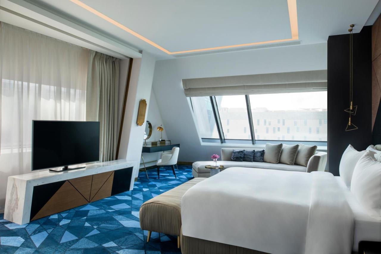 Steigenberger Hotel Doha Exterior photo The photo shows a modern hotel room featuring a sleek design. The room includes a large bed with white linens, a stylish seating area with gray and beige tones, and a flat-screen television mounted on a wall. There are large windows that allow natura