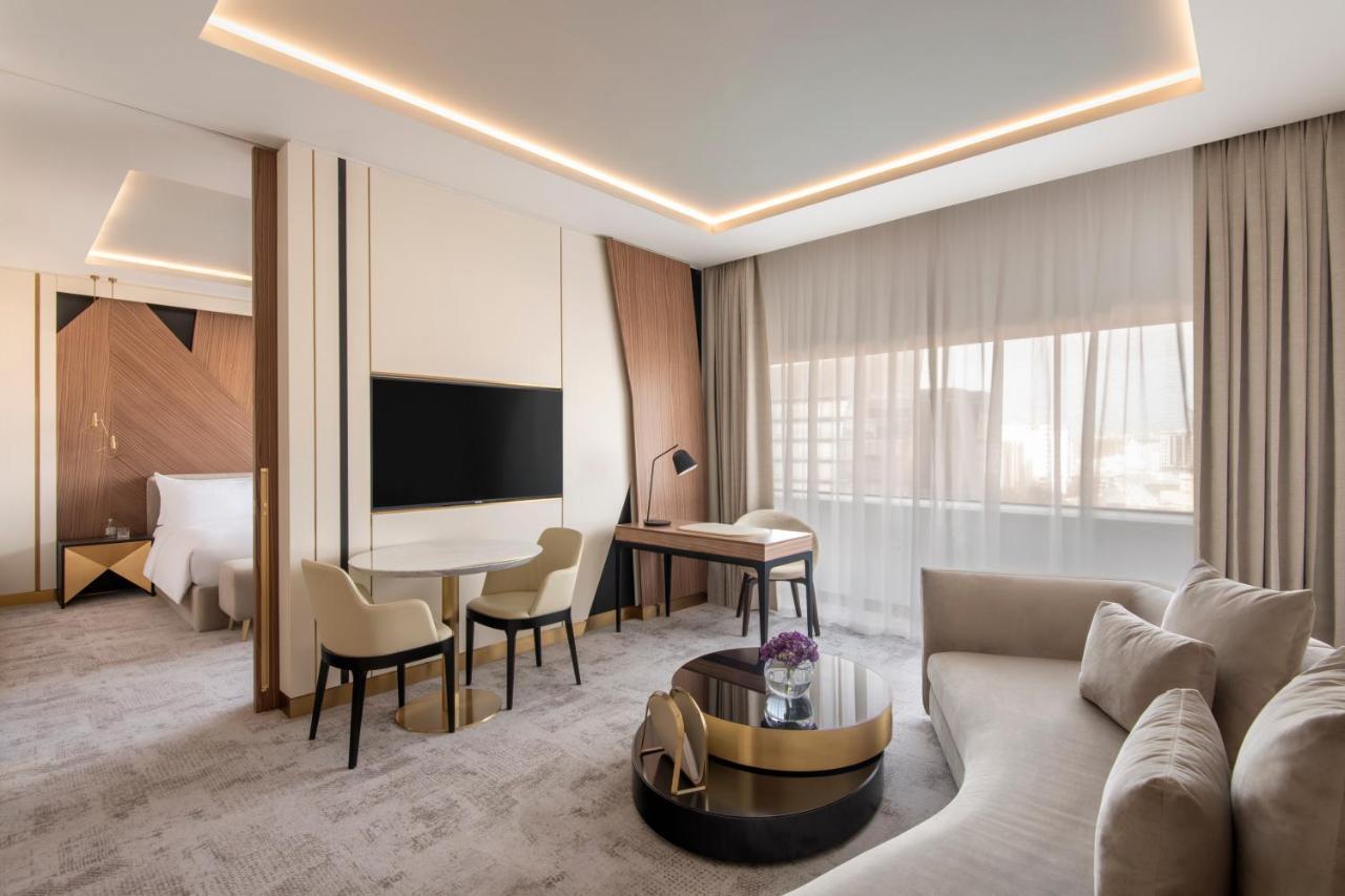 Steigenberger Hotel Doha Exterior photo The photo shows a modern hotel room that features a stylish and contemporary design. There is a comfortable seating area with a light-colored sofa and a round coffee table, along with two chairs and a small desk. A large flat-screen television is mou