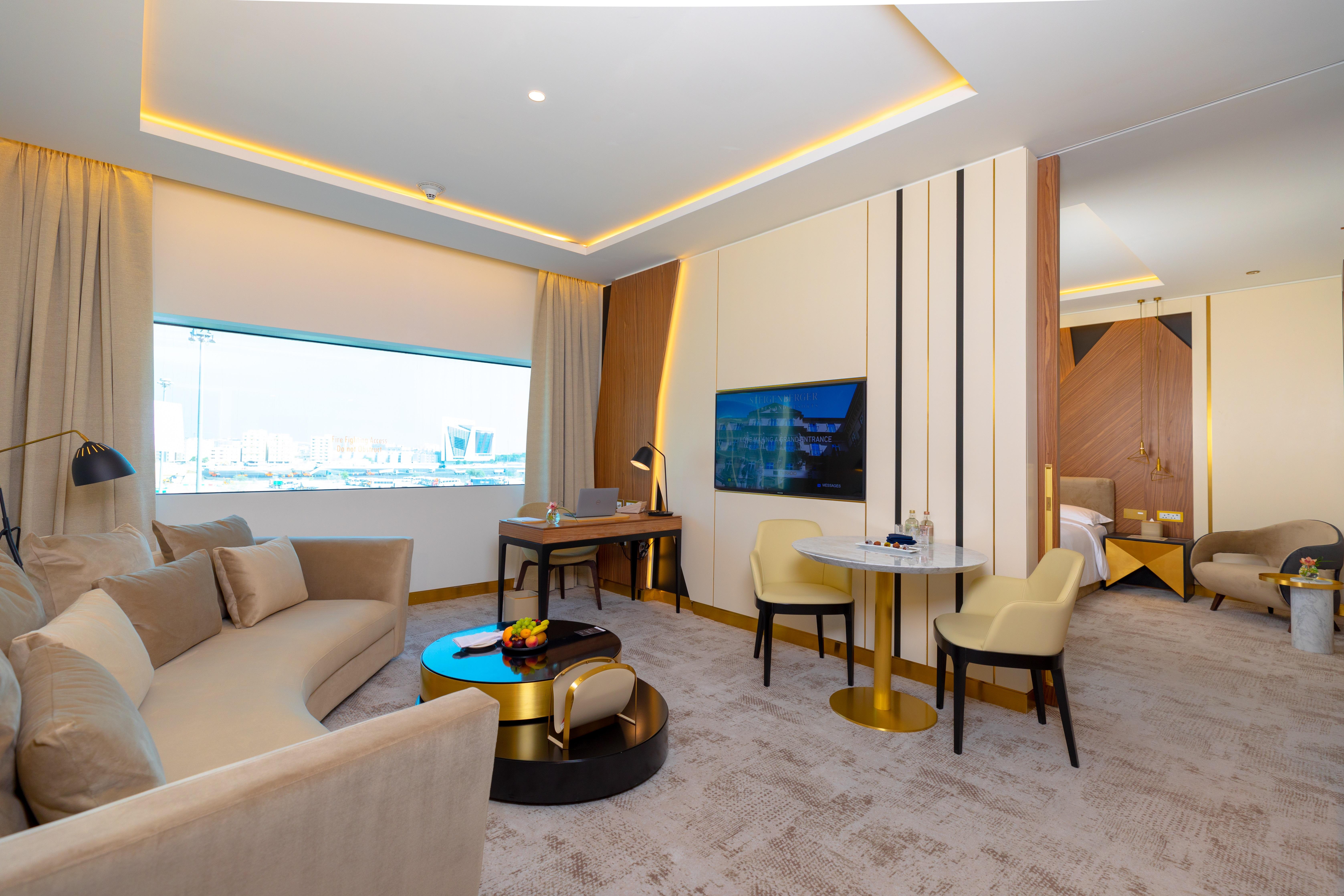 Steigenberger Hotel Doha Exterior photo The photo shows a modern and stylish hotel suite. It features a spacious living area with a curved sofa set and a small round table accompanied by two chairs. There is a large window that allows natural light to fill the room, enhancing its bright an
