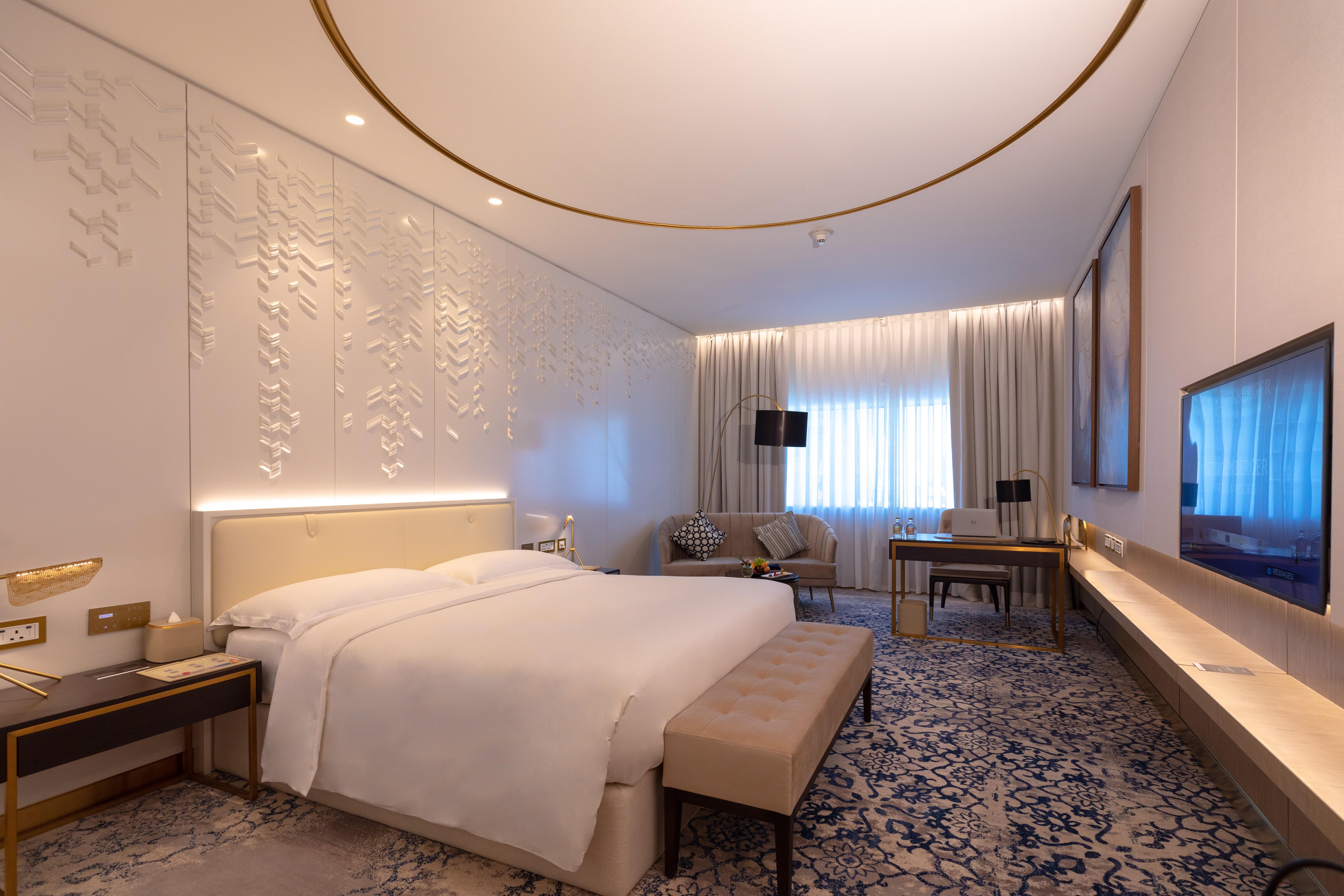 Steigenberger Hotel Doha Exterior photo The photo shows a modern hotel room designed with a contemporary aesthetic. It features a large bed with white linens, a soft beige bench at the foot of the bed, and elegant furnishings including a bedside table and a comfortable seating area with ch