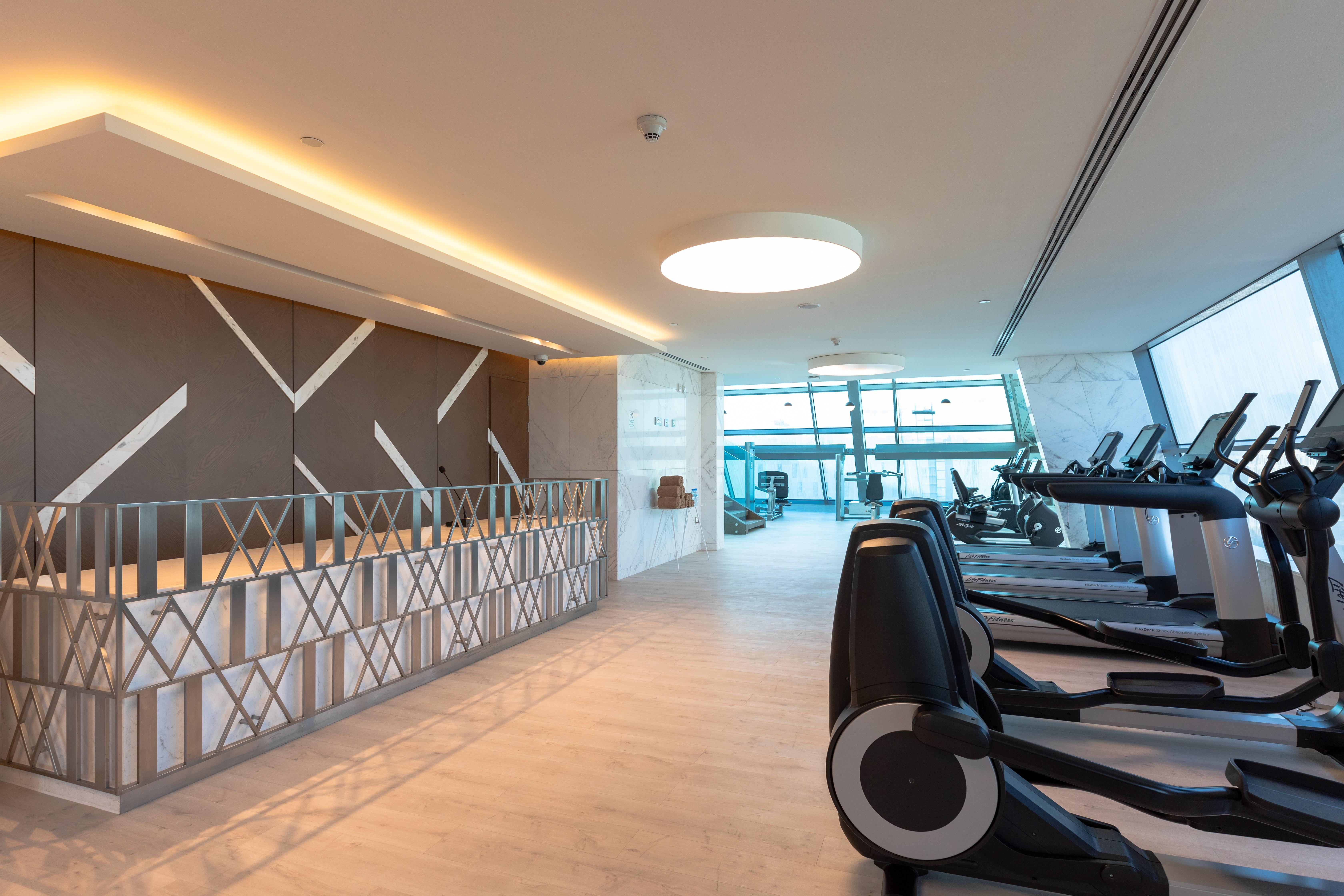 Steigenberger Hotel Doha Exterior photo The photo shows the interior of a modern gym. On the left side, there is a reception area with a stylish design featuring geometric patterns on the wall. The area is well-lit with natural light coming in from large windows, providing a view outside. 