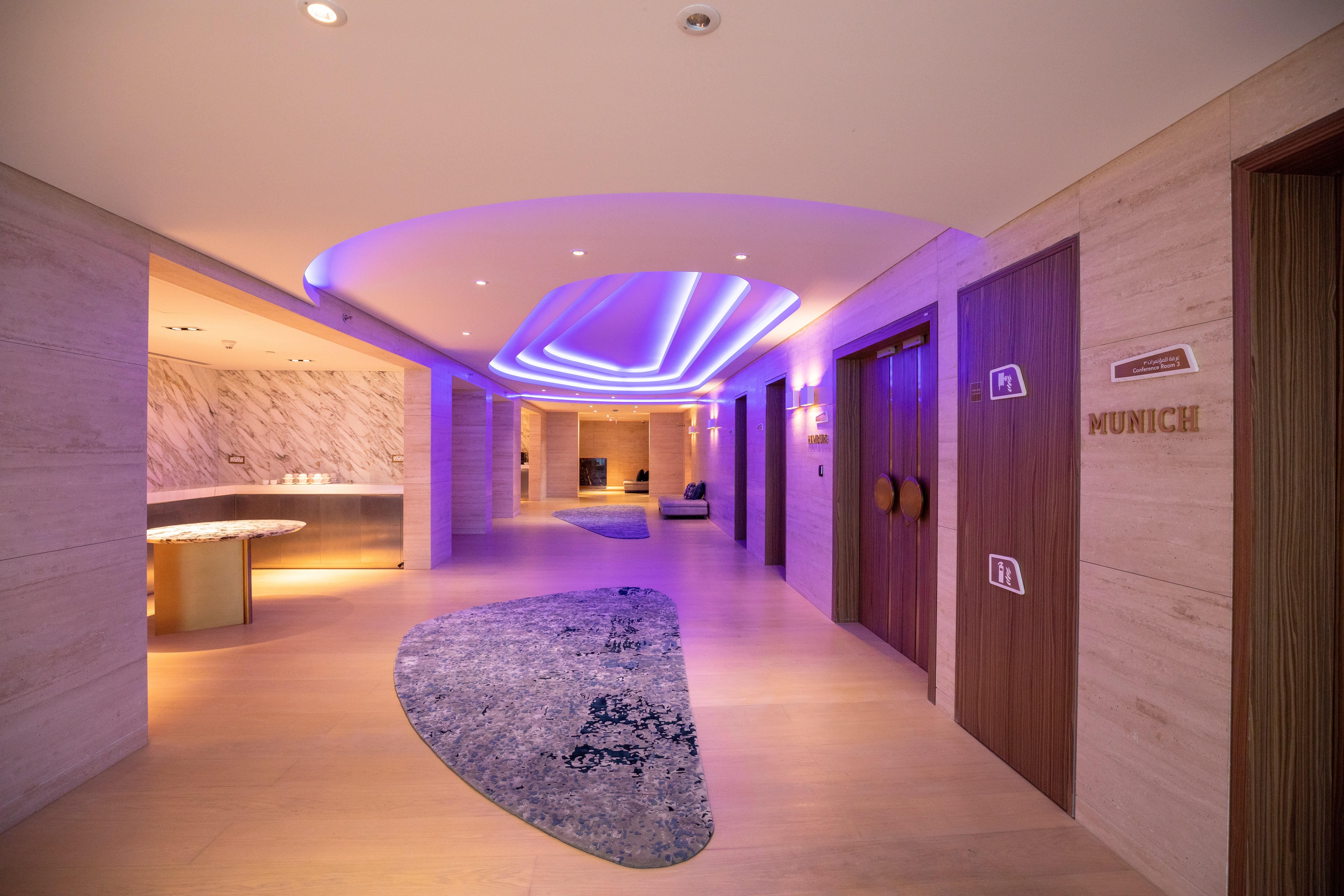 Steigenberger Hotel Doha Exterior photo The photo shows a modern hallway in a stylish setting. The corridor features soft, ambient lighting with a purple hue, creating a calming atmosphere. On the left side, there is a curved reception area with a smooth surface. The floor is decorated wit