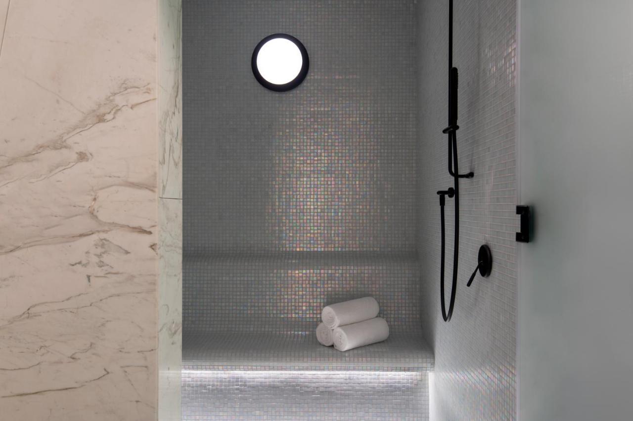Steigenberger Hotel Doha Exterior photo The photo features a modern shower space. The walls are adorned with small mosaic tiles that create a sleek and reflective surface. There is a round window that allows natural light to enter, enhancing the ambiance. A black shower head and faucet are