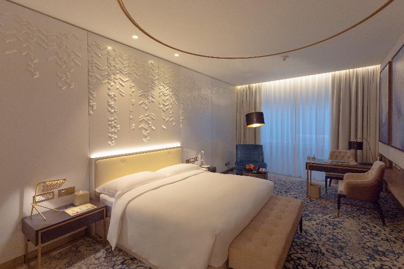 Steigenberger Hotel Doha Exterior photo The photo shows a modern hotel room featuring a contemporary design. The room includes a large bed with a padded headboard, positioned against a textured wall with decorative patterns. To the side, there is a stylish bedside table with a lamp. A comf